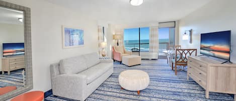 A modern hotel room with a large window offering a view of the ocean, a grey sofa, a small round ottoman, a wooden dresser, and a flat-screen TV.