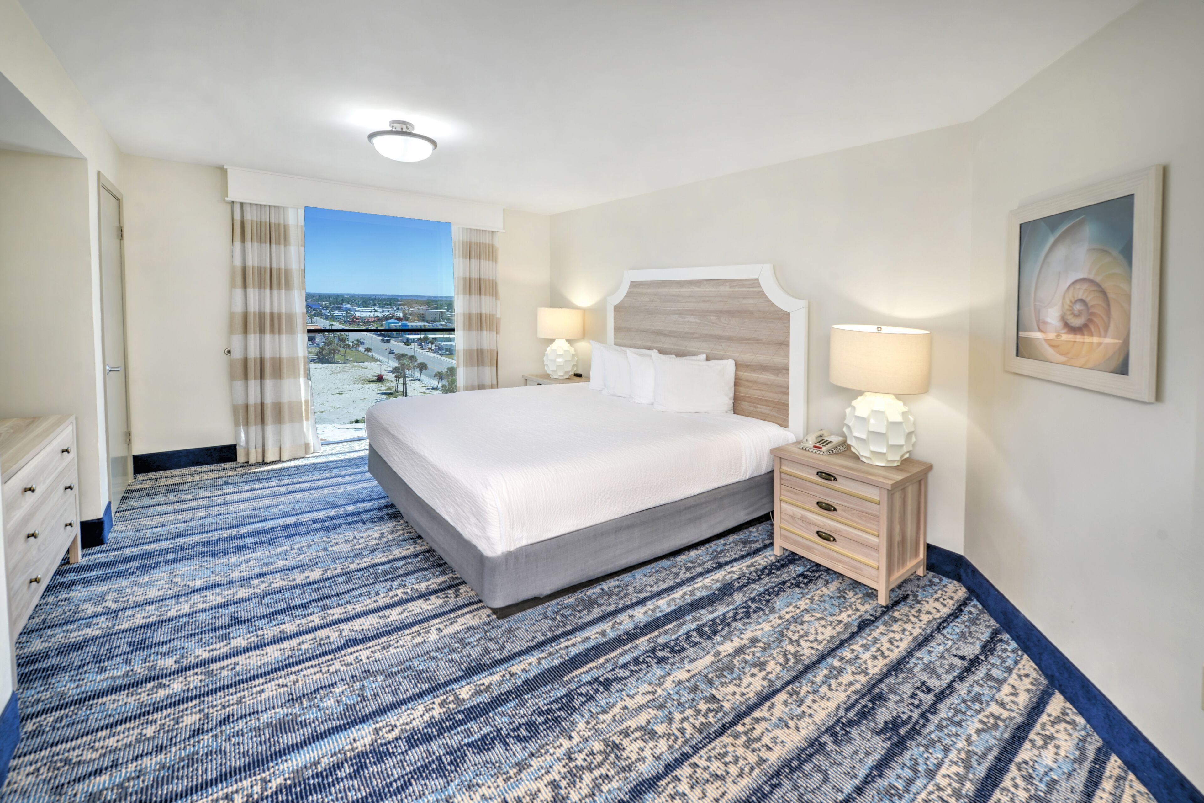 A hotel room with a large bed, two bedside tables with lamps, a view of the city, and a balcony.