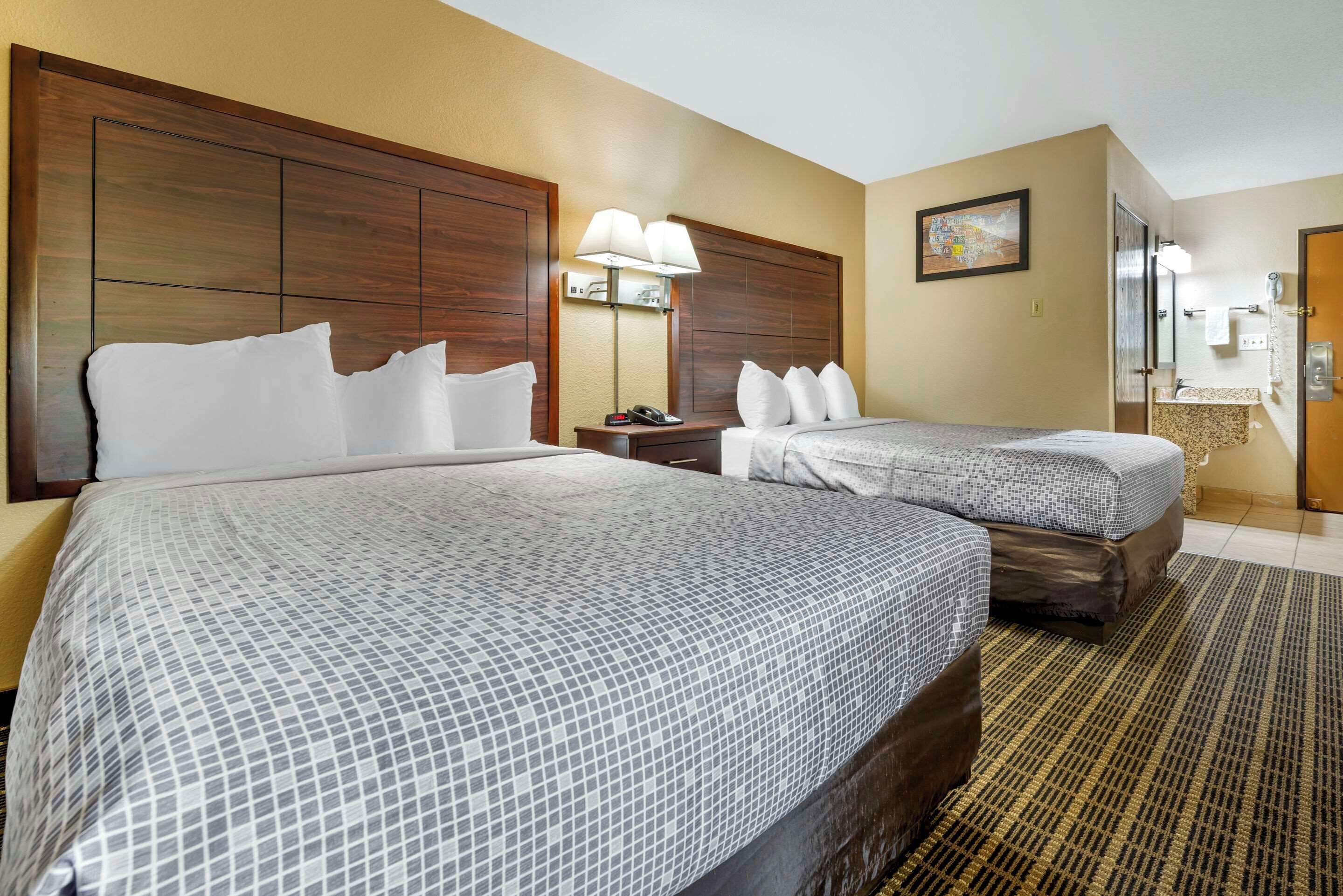 A hotel room with two beds, a large wooden headboard, a nightstand with a lamp, a bathroom with a sink and mirror, and a painting on the wall.