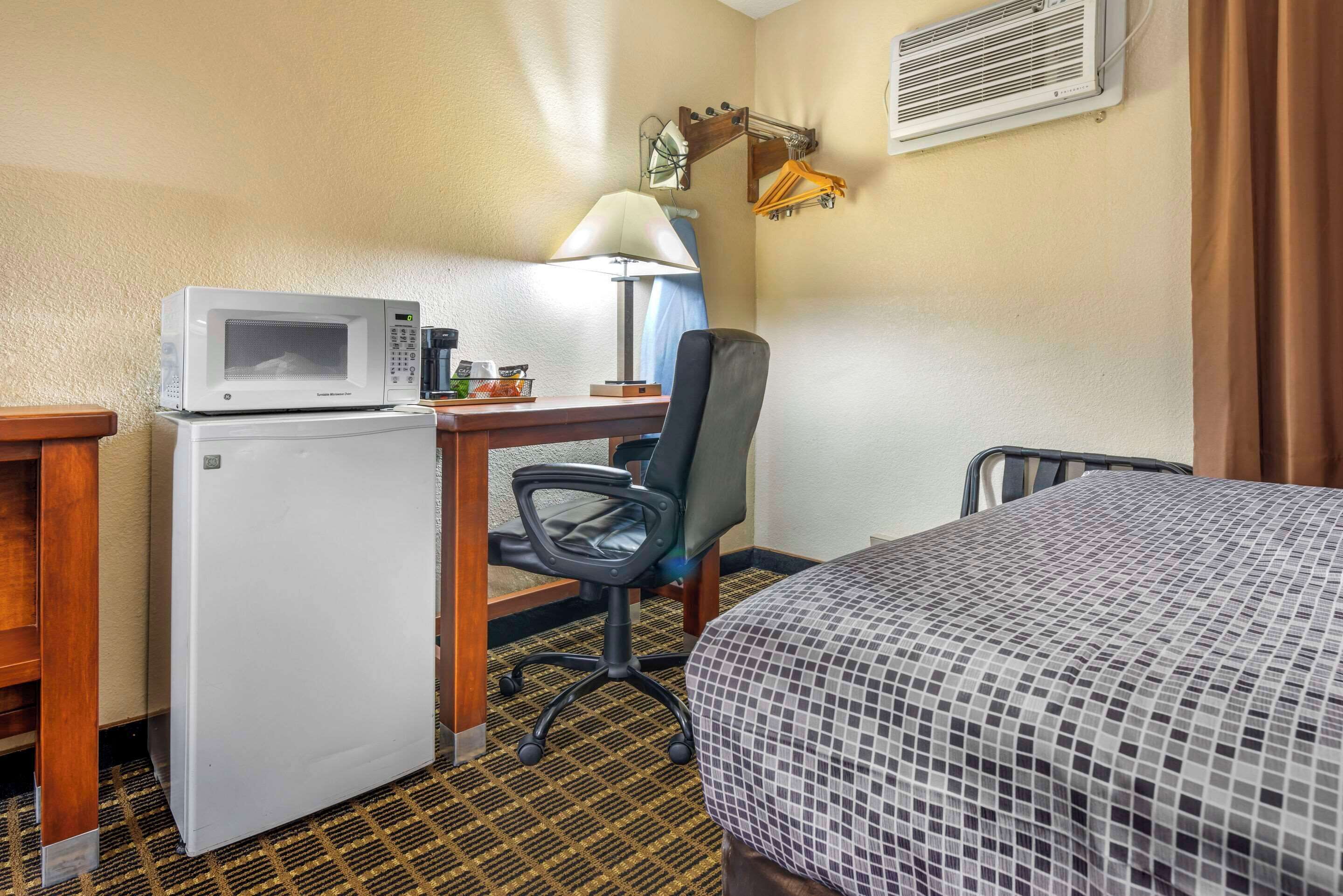 A compact hotel room with a kitchenette, a desk with a lamp, a chair, and a bed with a patterned bedspread.