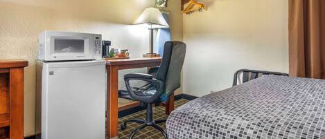 A compact hotel room with a kitchenette, a desk with a lamp, a chair, and a bed with a patterned bedspread.