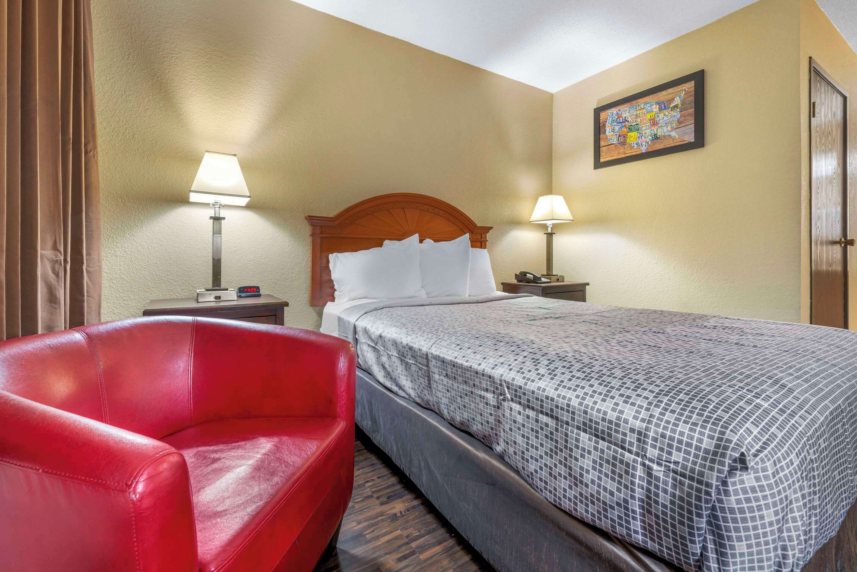 A hotel room with a bed, two bedside lamps, a red chair, a wooden headboard, and a wall map.