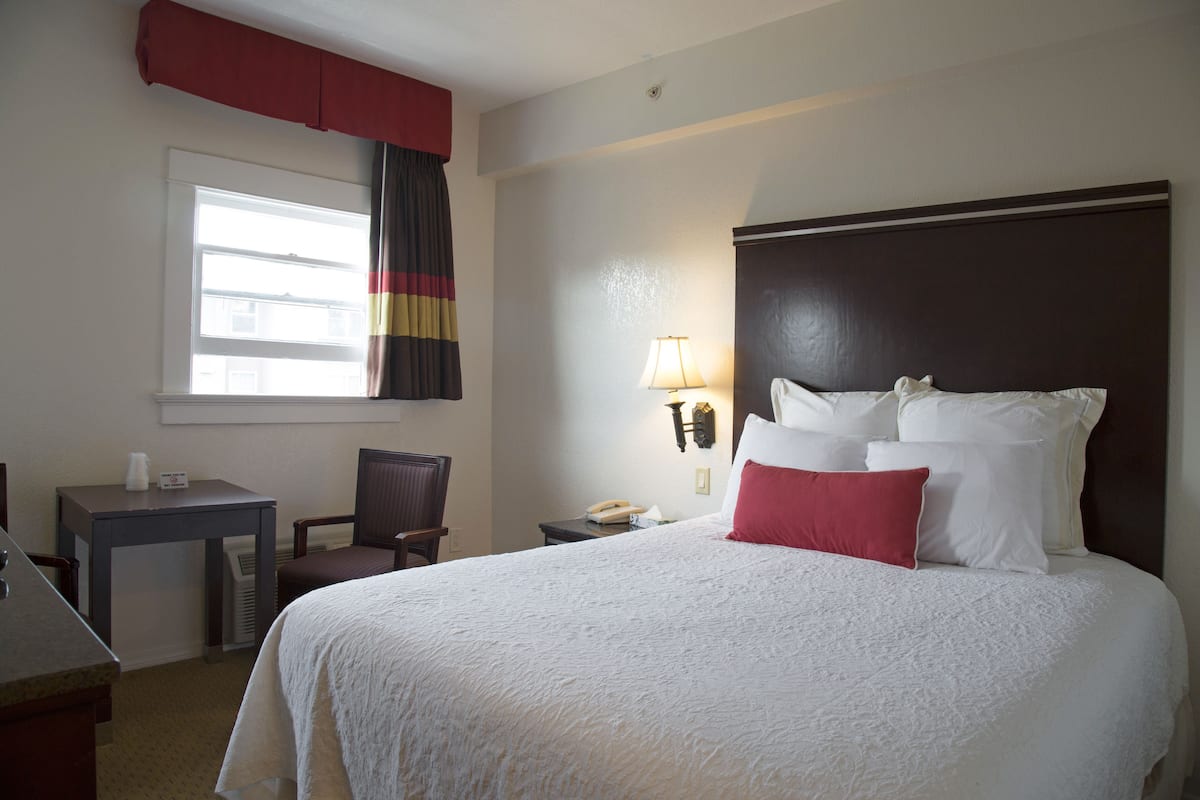 Una habitación de hotel con una cama grande, un escritorio, una silla, una ventana con cortinas y una lámpara de pared.