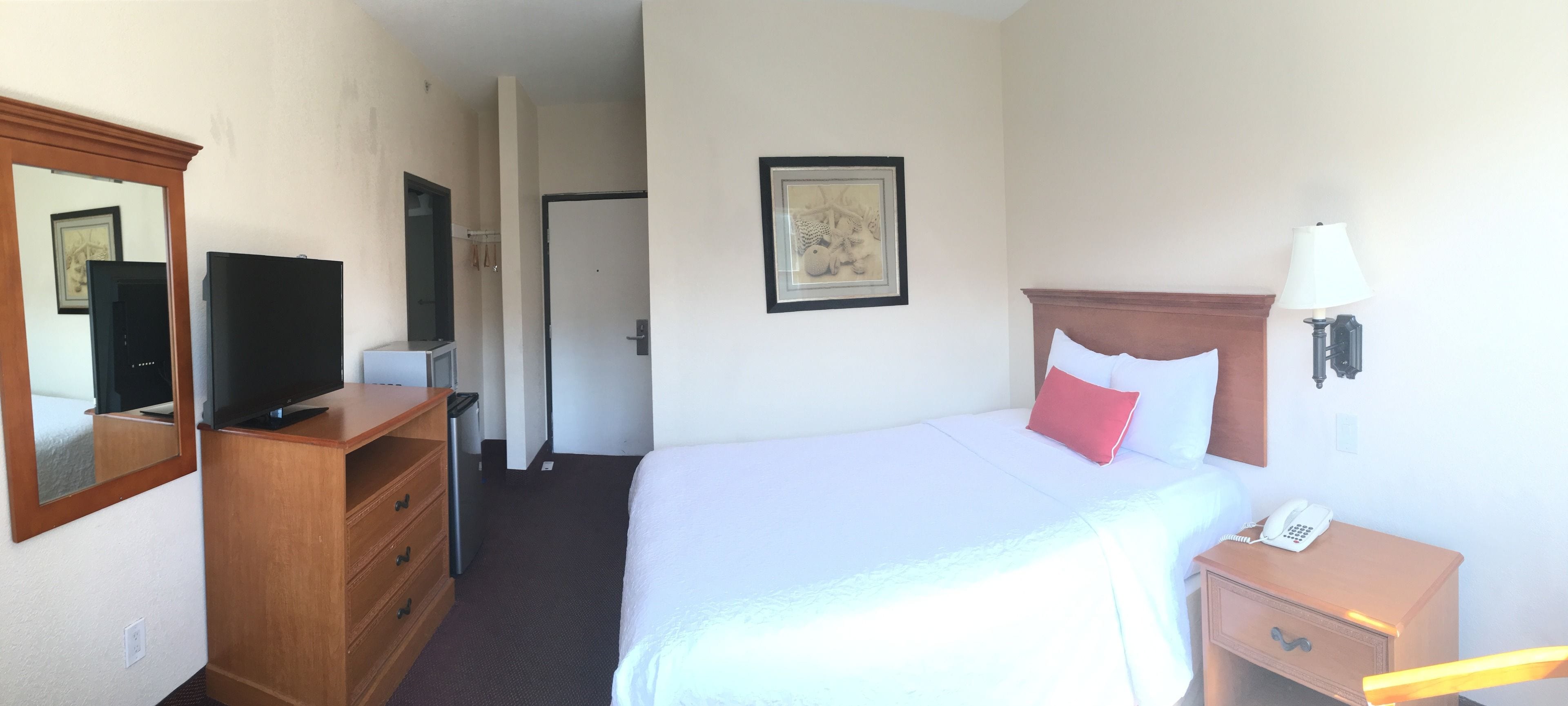 Ein Hotelzimmer mit einem Bett, einem Nachttisch mit Lampe, einem Telefon, einem Schminktisch, einem Fernseher und einem gerahmten Bild an der Wand.