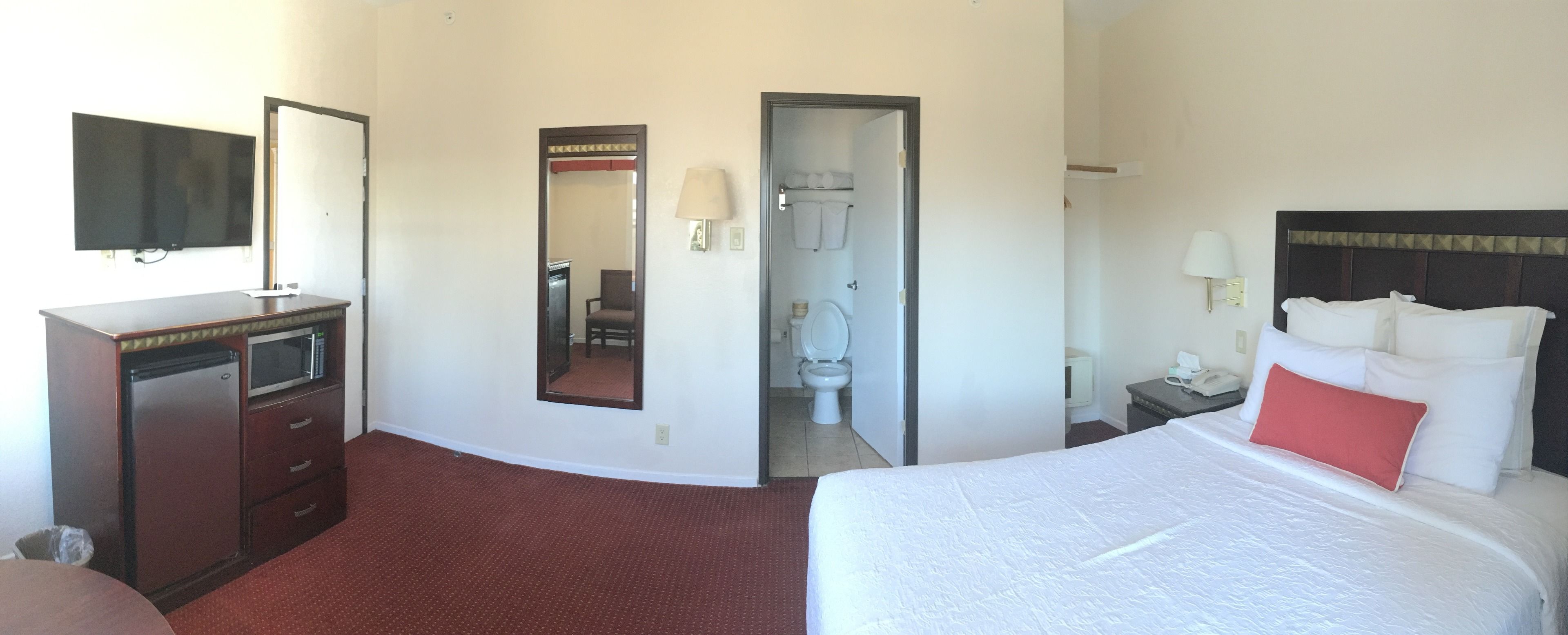A hotel room with a bed, a desk, a television, a mirror, and a bathroom visible through an open door.