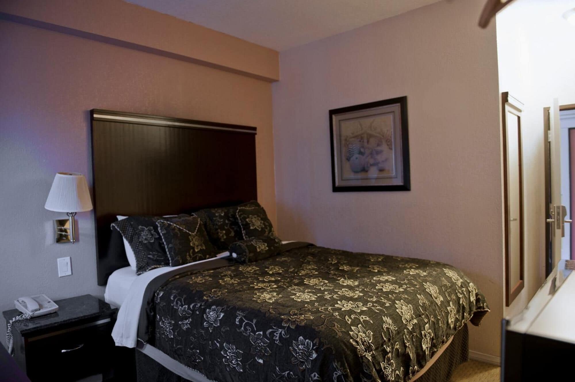 A hotel room with a bed, a nightstand, a lamp, a framed picture on the wall, and a mirror.