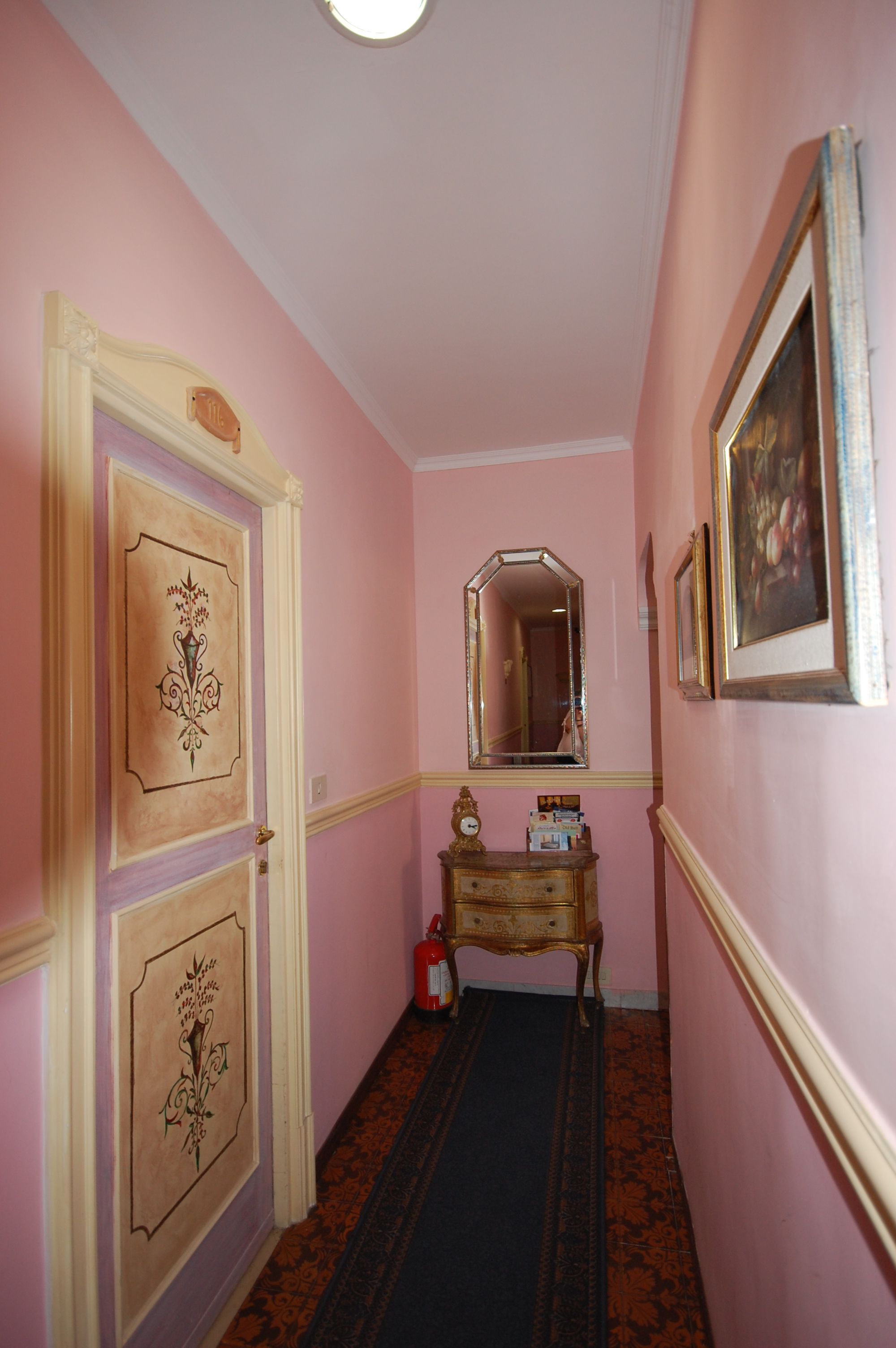 A hallway with pink walls, a wooden door with a floral design, a mirror, a small wooden dresser, and a patterned carpet.