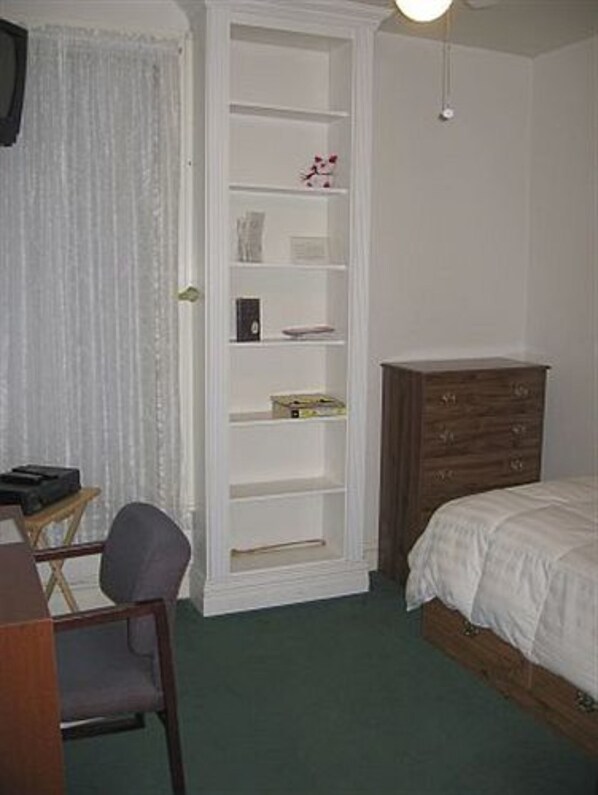 Une chambre à coucher comprenant un lit, une commode en bois, une étagère blanche, un bureau avec une chaise et une fenêtre avec des rideaux.