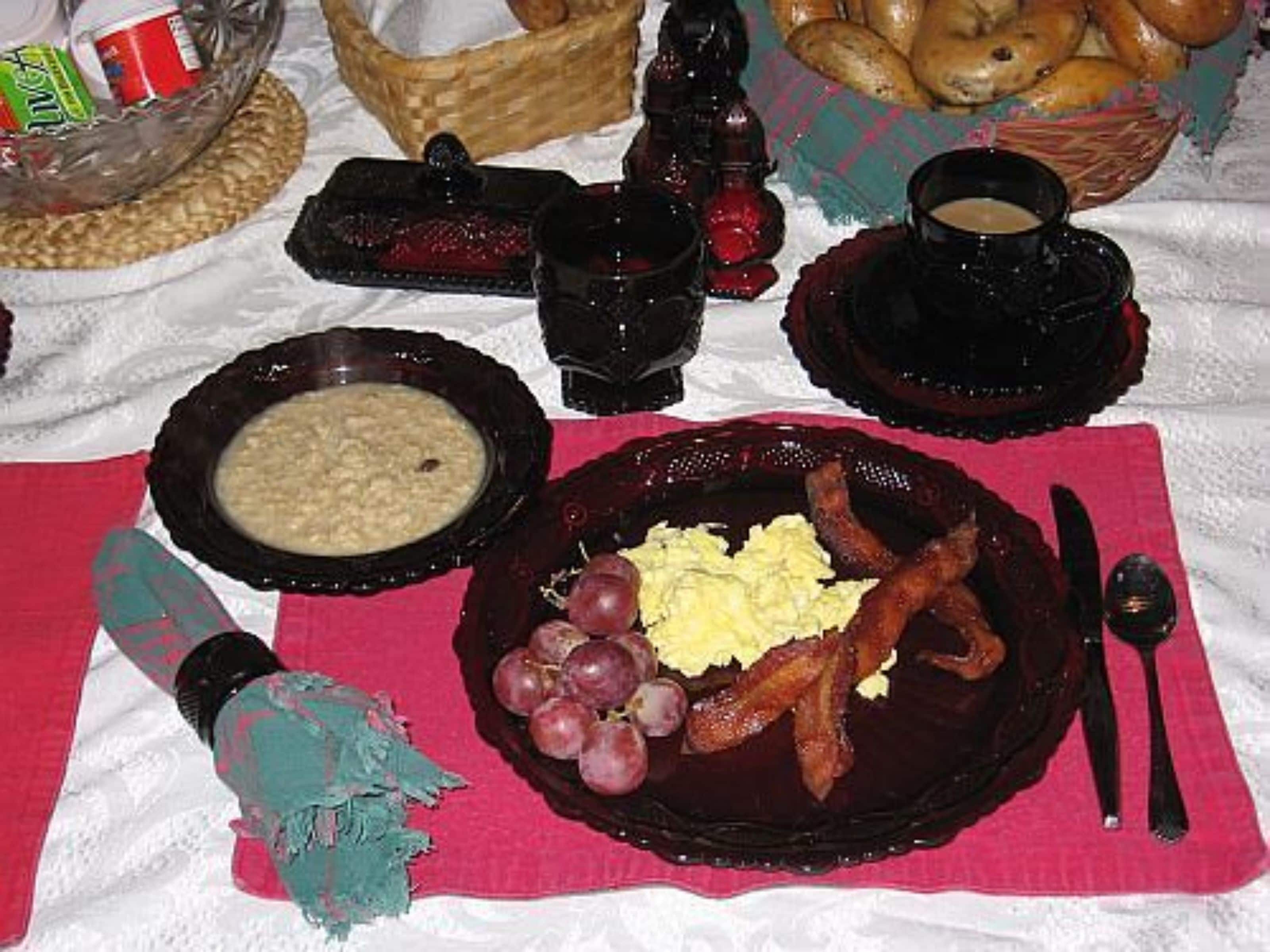 A breakfast setting with a plate of eggs, bacon, and grapes, a bowl of soup, and a basket of bread.
