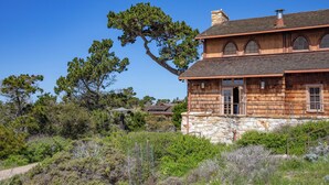 A two-story house with a stone foundation and wooden upper structure, surrounded by dense vegetation and a clear blue sky.