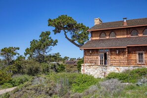 A two-story house with a stone foundation and wooden upper structure, surrounded by dense vegetation and a clear blue sky.