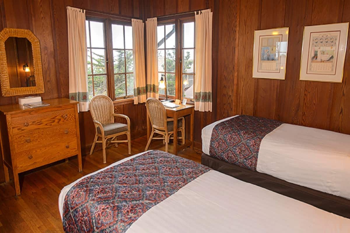 A room with wooden walls, two beds with patterned bedspreads, a wooden dresser, a chair, a desk, and a window with curtains.