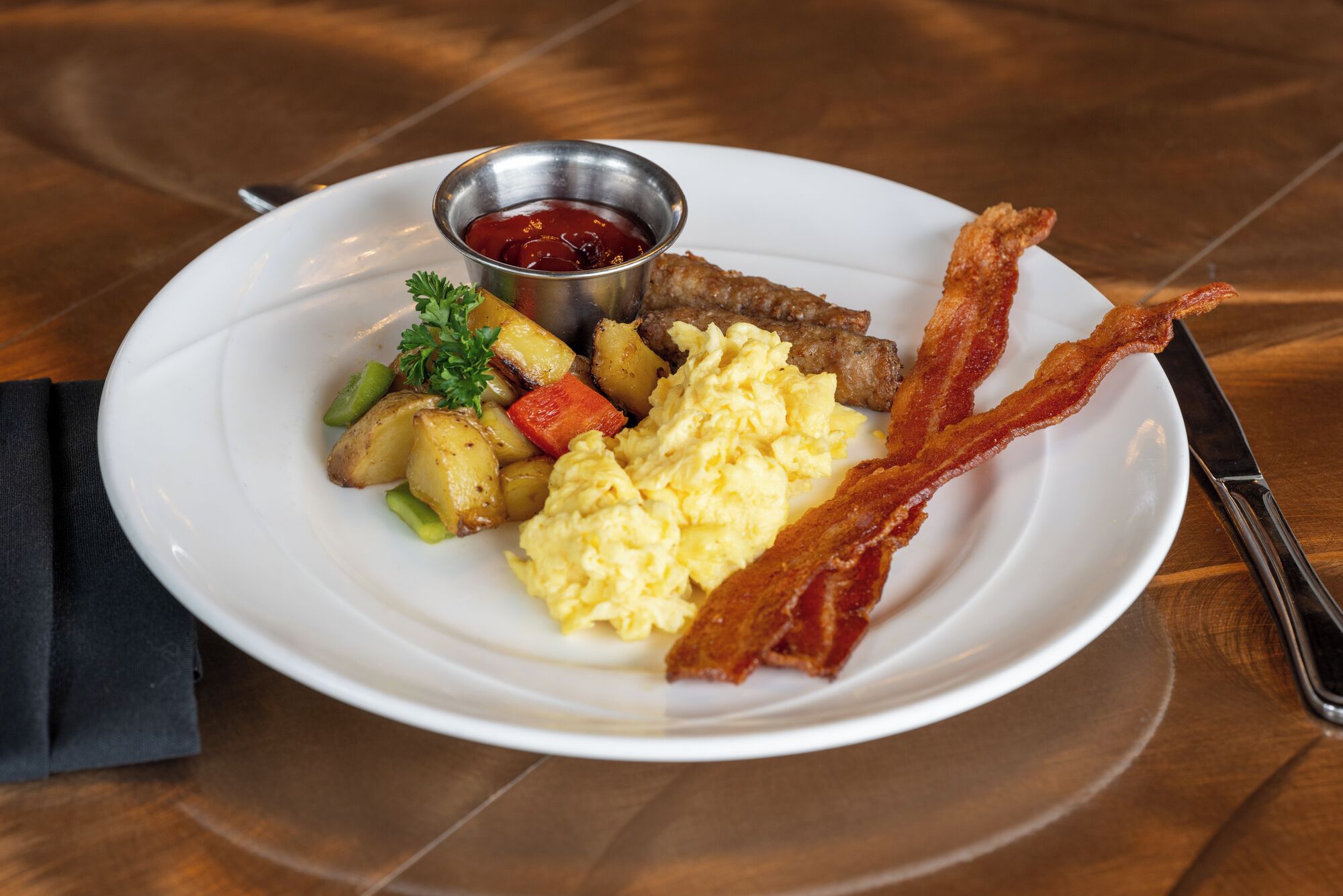 A plate of breakfast food including scrambled eggs, bacon, potatoes, and a side of ketchup.