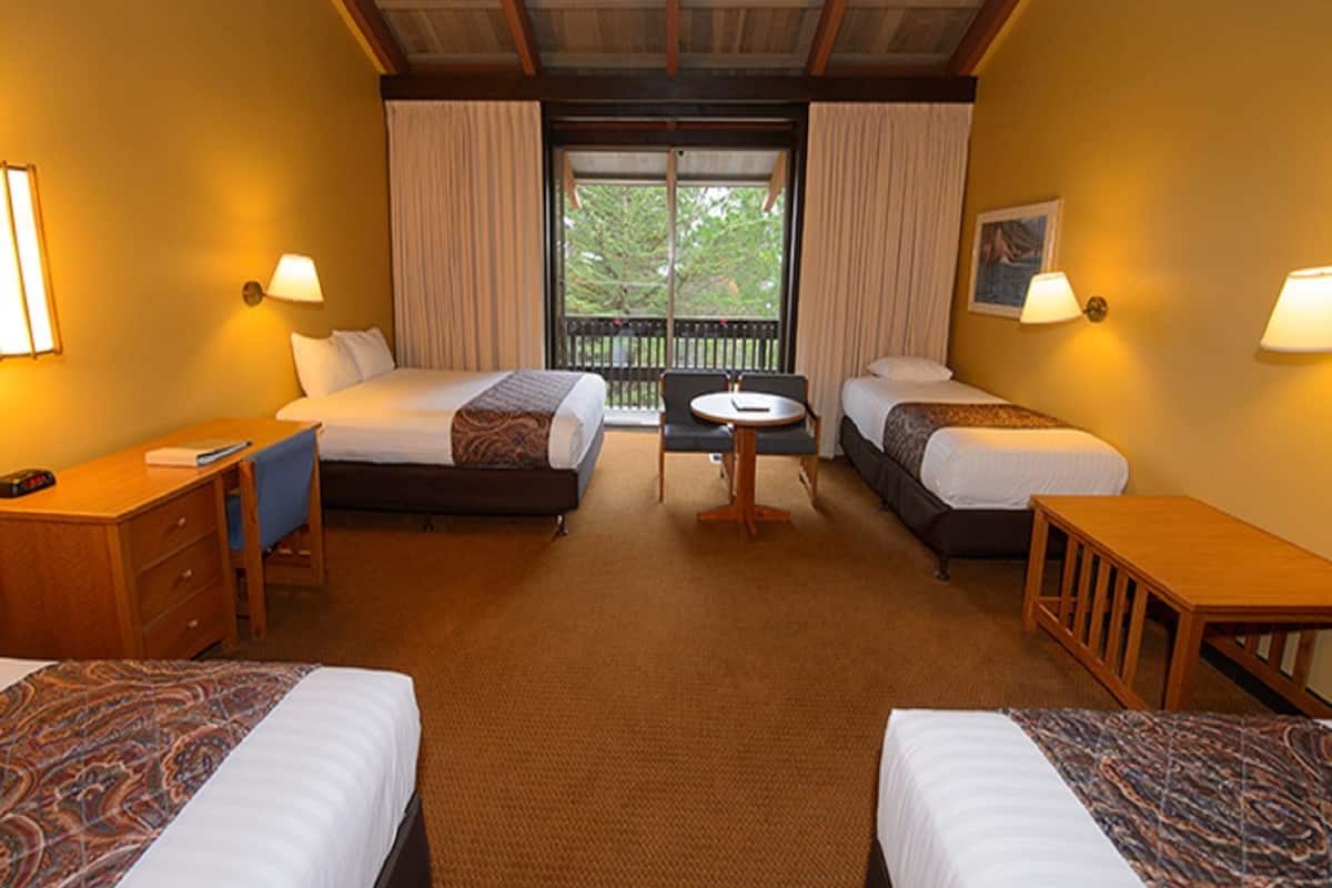 A hotel room with two beds, a desk, a chair, a small table, and a balcony with a view of trees.