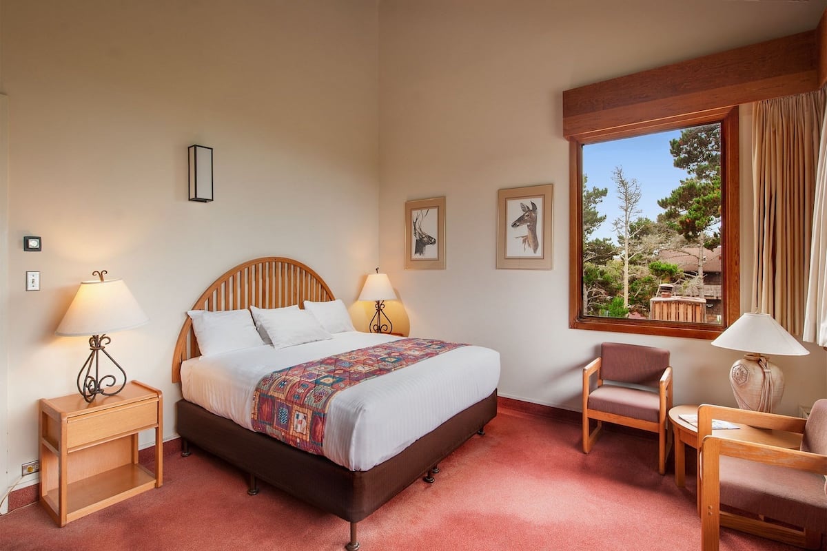 A bedroom with a wooden bed, two bedside tables, a chair, a lamp, and a window with a view of trees.