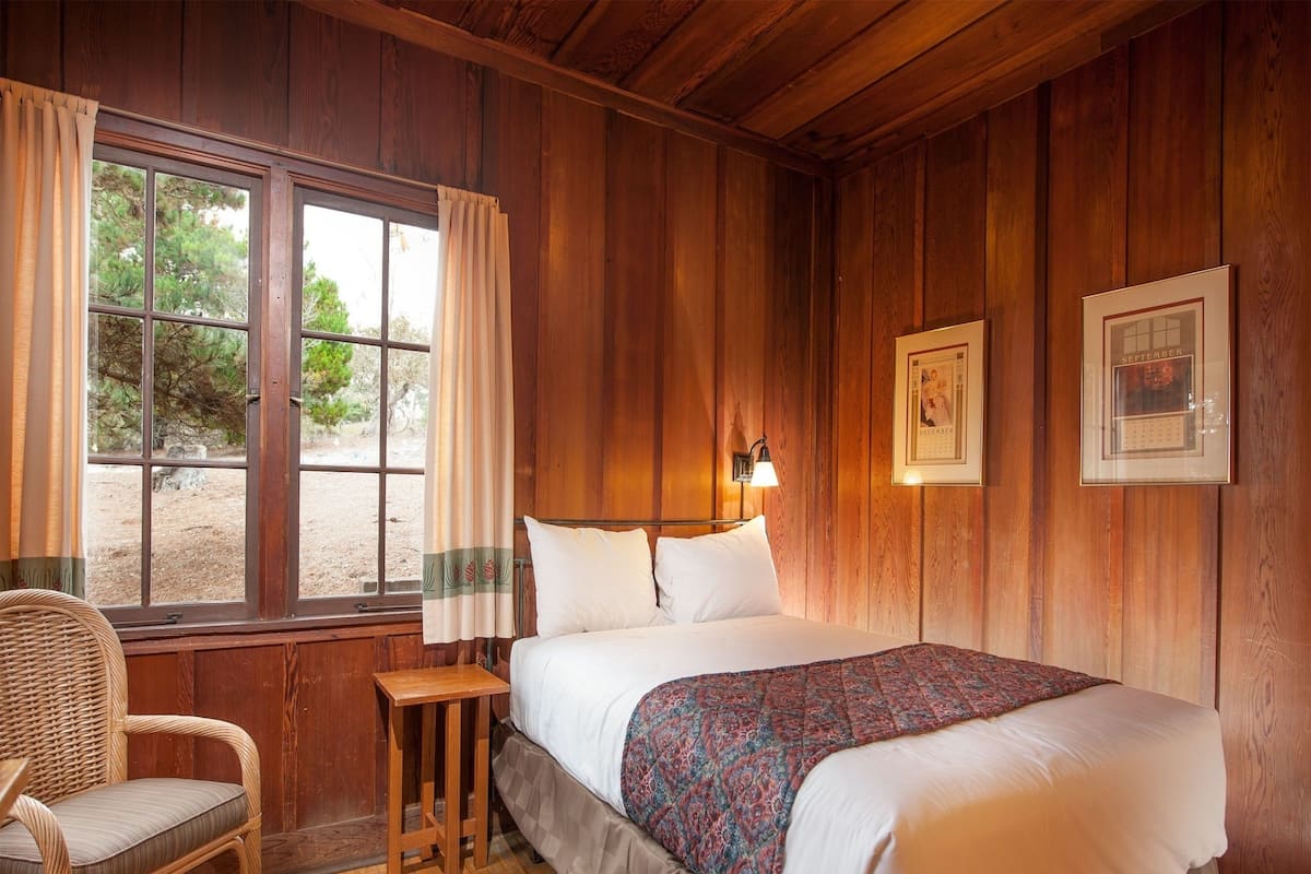 A wooden room with a bed, a chair, a small table, and a window with curtains.