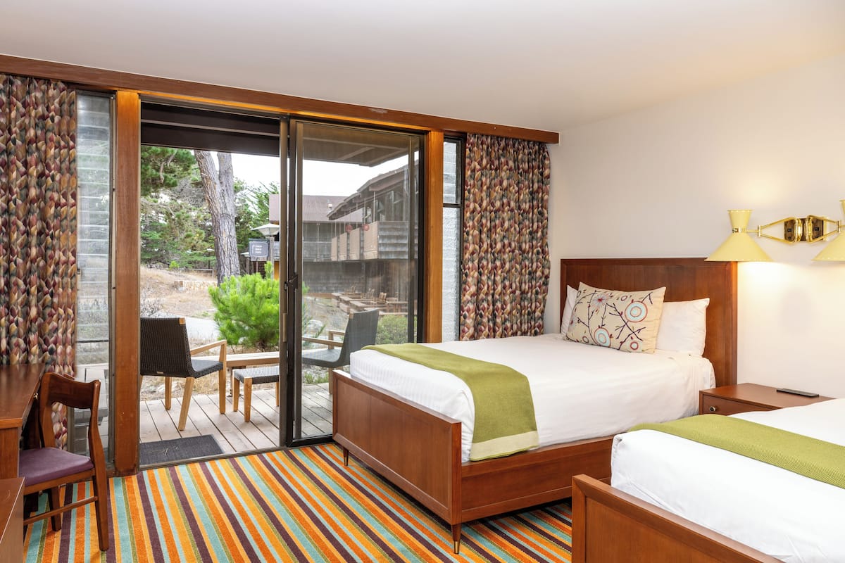 A hotel room with a large bed, a desk, and a chair. There is a sliding glass door leading to an outdoor area with a patio and a view of a building.