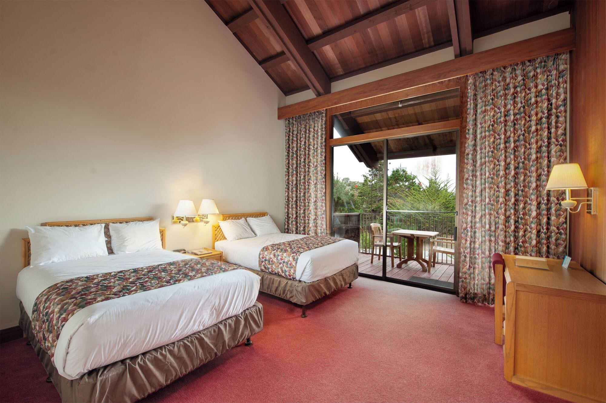 A hotel room with two beds, a wooden ceiling, a balcony with a table and chairs, and a small wooden desk with a lamp.