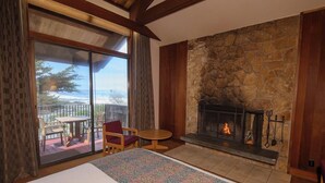 A room with a large bed, a stone fireplace, and a balcony with a view of the ocean.