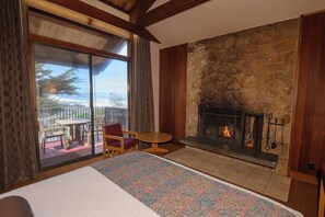 A room with a large bed, a stone fireplace, and a balcony with a view of the ocean.