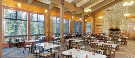 A spacious dining area with high ceilings, large windows, and a stone fireplace. Tables are neatly arranged with chairs, and the floor has a herringbone pattern.