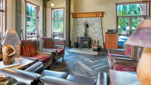 A cozy living room with leather sofas, a stone fireplace, and large windows offering a view of the outdoors.