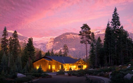 A lodge with a wooden exterior, surrounded by evergreen trees and snow-capped mountains.