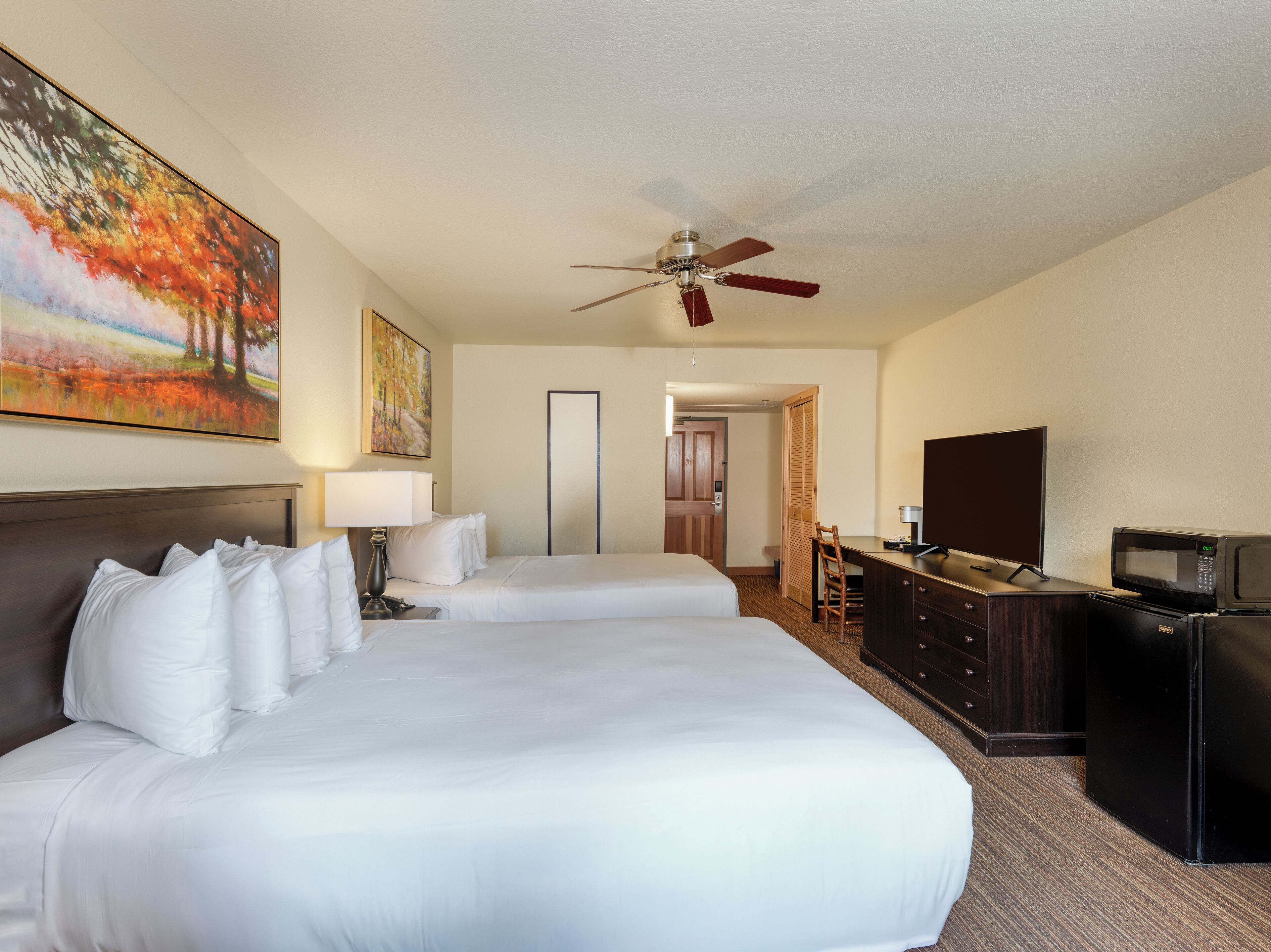 A hotel room with two beds, a desk with a TV and microwave, a ceiling fan, and artwork on the walls.