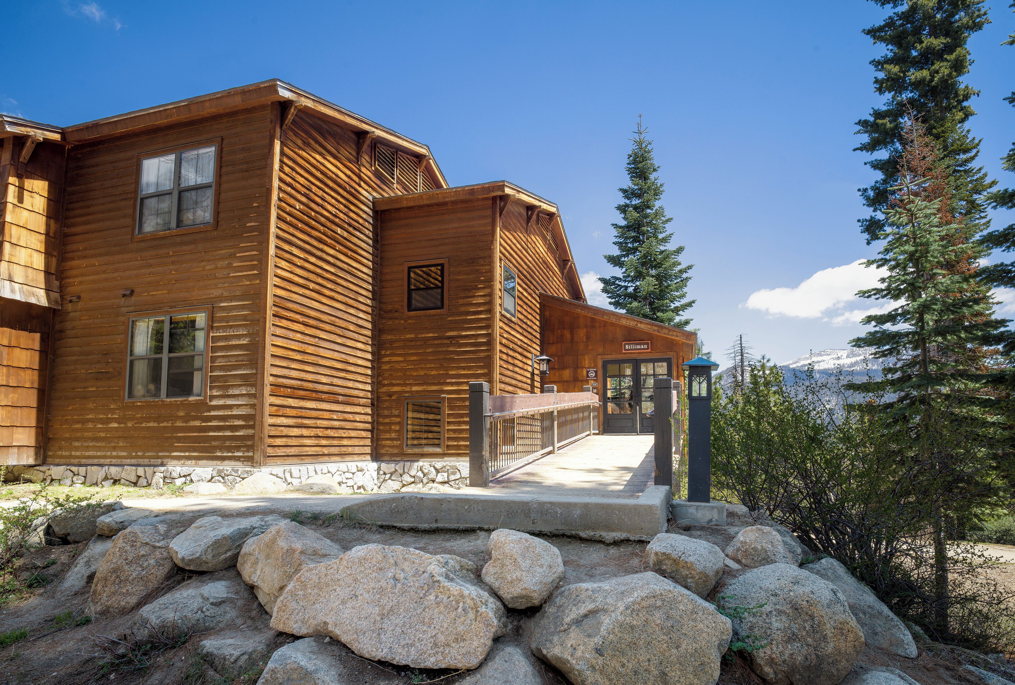 A wooden cabin with a stone foundation, large windows, and a stone pathway leading to the entrance.