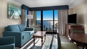 Una camera d'albergo con divano, tavolino, televisione, balcone vista mare e un quadro appeso al muro.