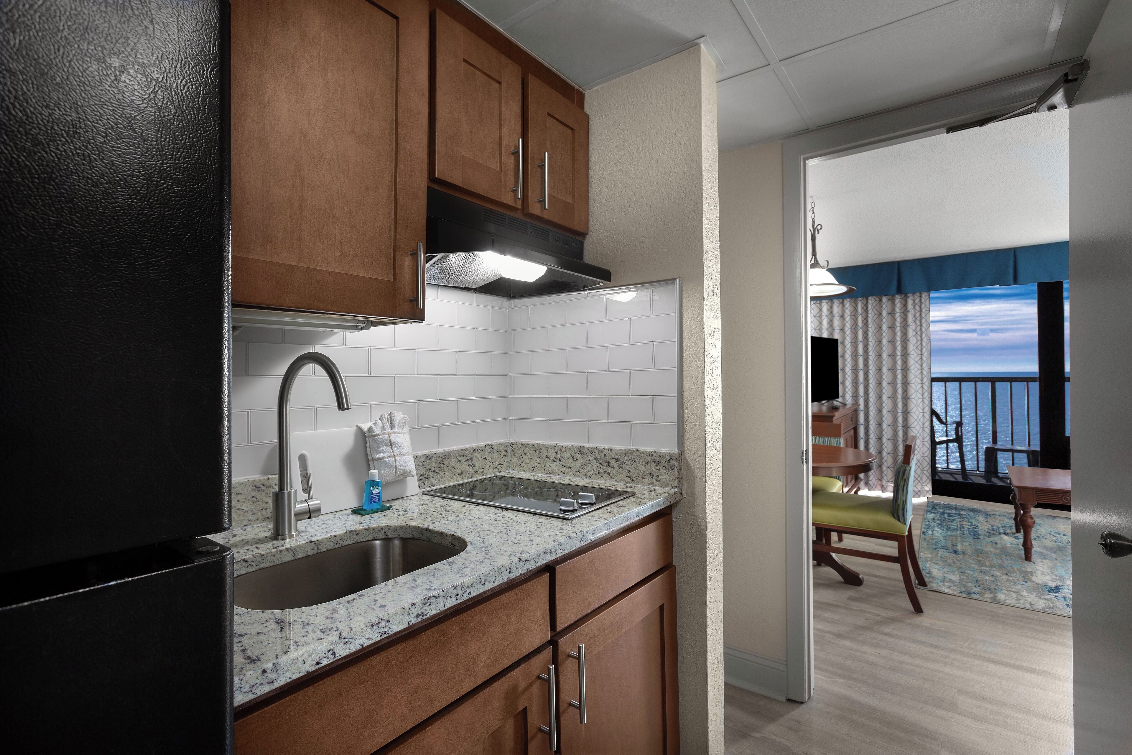 A compact kitchen with a sink, stove, and wooden cabinets. A dining area with a table and chairs is visible through an open door.