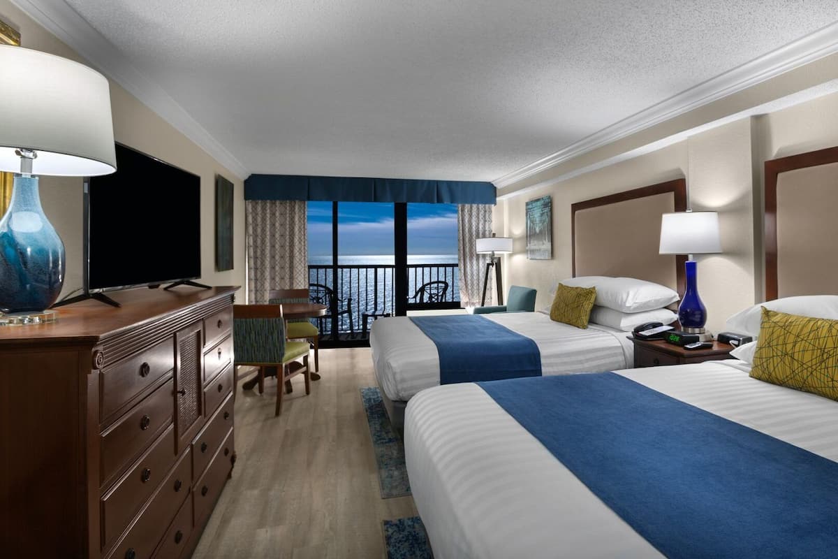 A hotel room with two beds, a desk, a TV, and a balcony with a view.