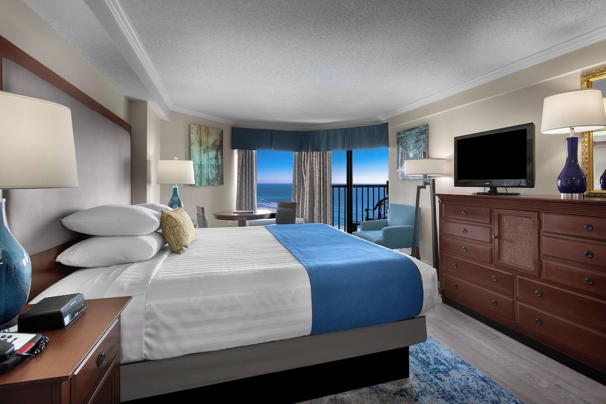 A hotel room with a bed, a TV, a dresser, and a view of the ocean.