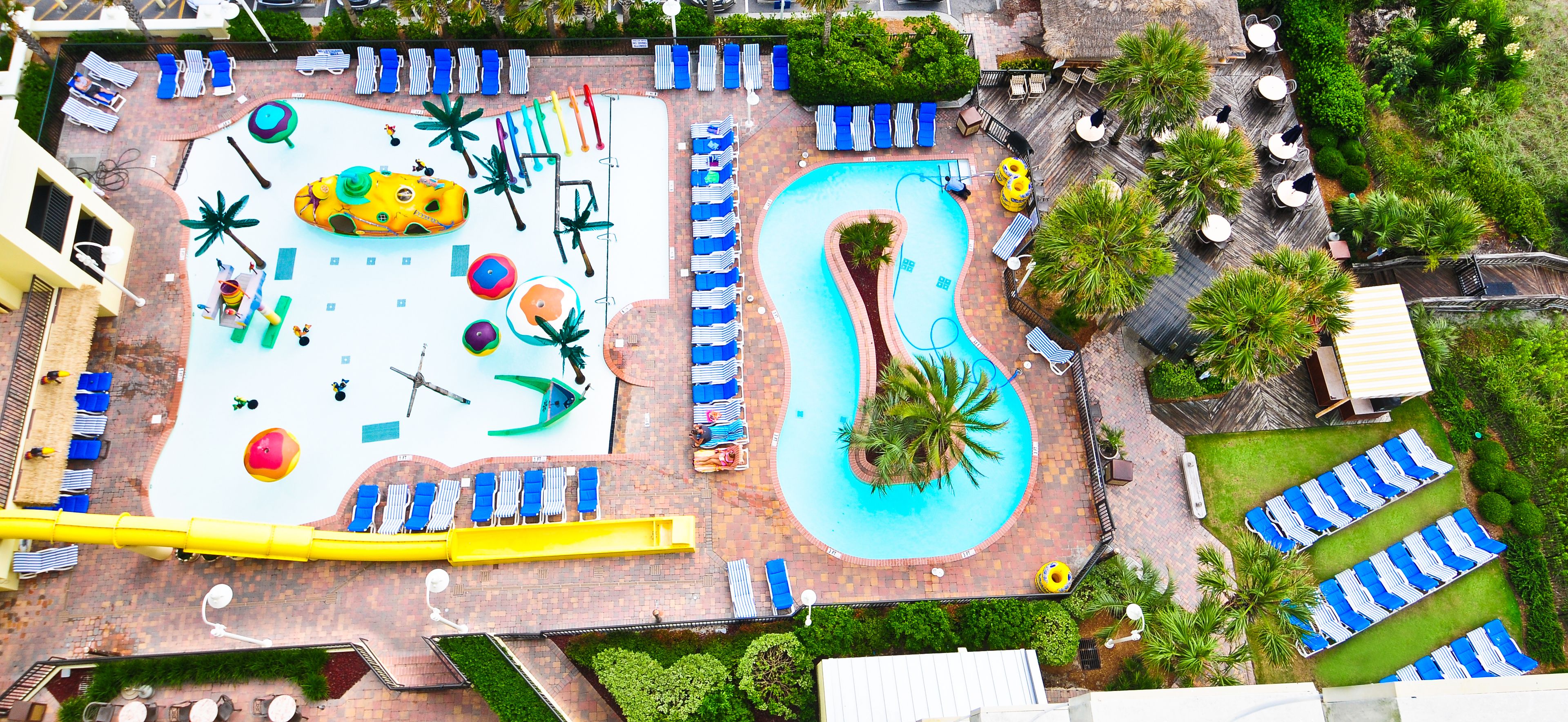 An aerial view of a resort with a large pool, multiple lounge areas with chairs and umbrellas, and a slide.