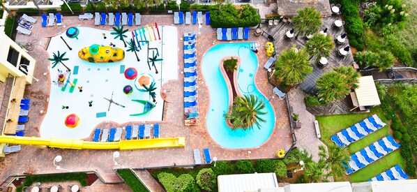 An aerial view of a resort with a large pool, multiple lounge areas with chairs and umbrellas, and a slide.