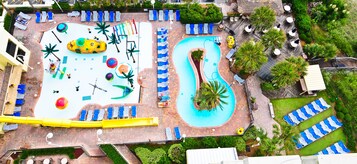 An aerial view of a resort with a large pool, multiple lounge areas with chairs and umbrellas, and a slide.