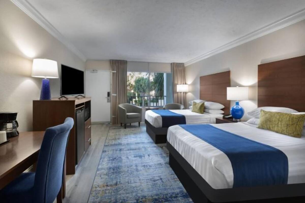 A hotel room with two beds, a desk with a chair, a television, and a view of the outdoors.