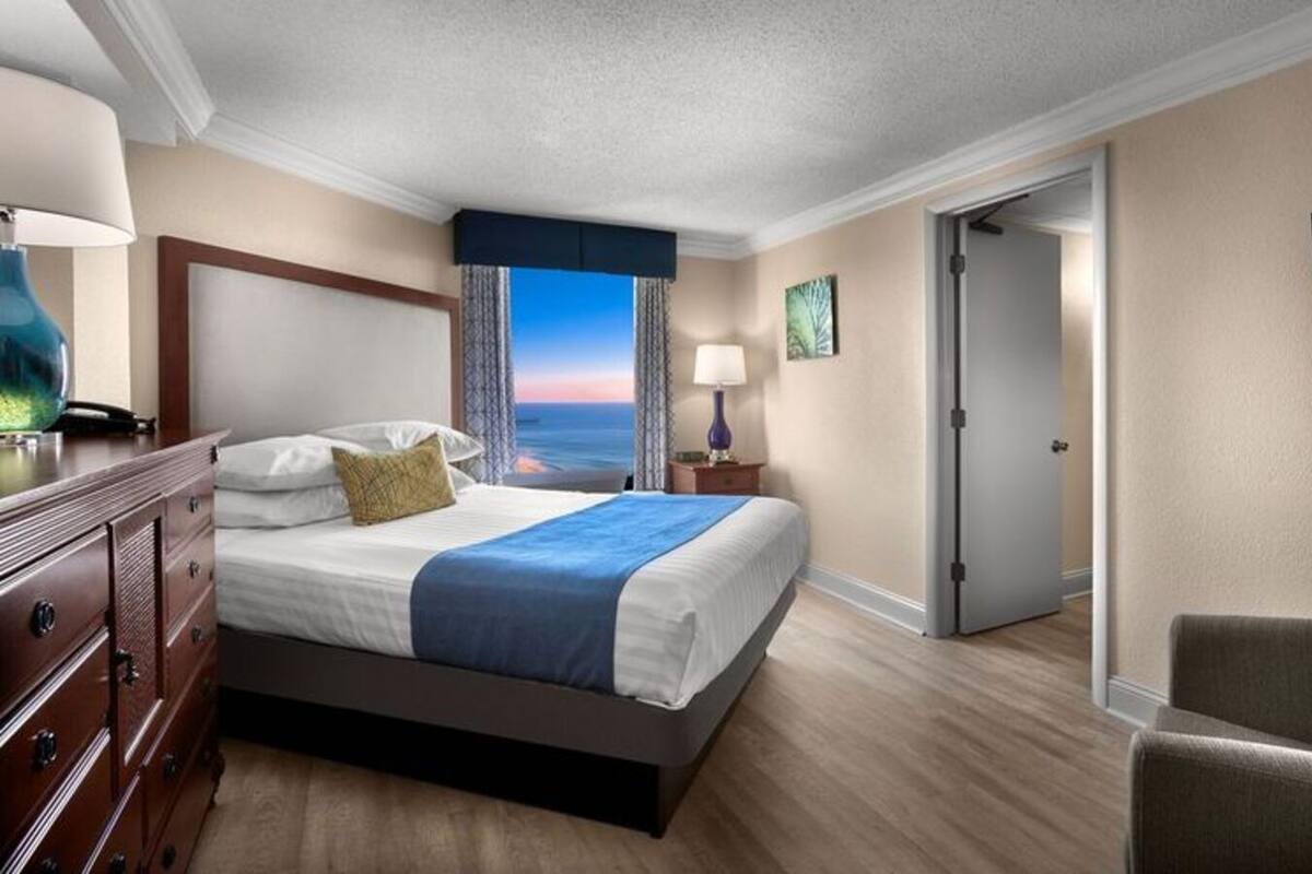 A hotel room with a bed, a desk with a lamp, a chair, a door, and a view of the ocean.