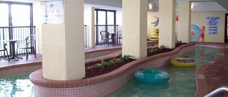 An indoor swimming pool with steps leading into the water, surrounded by seating areas and a lifeguard station.