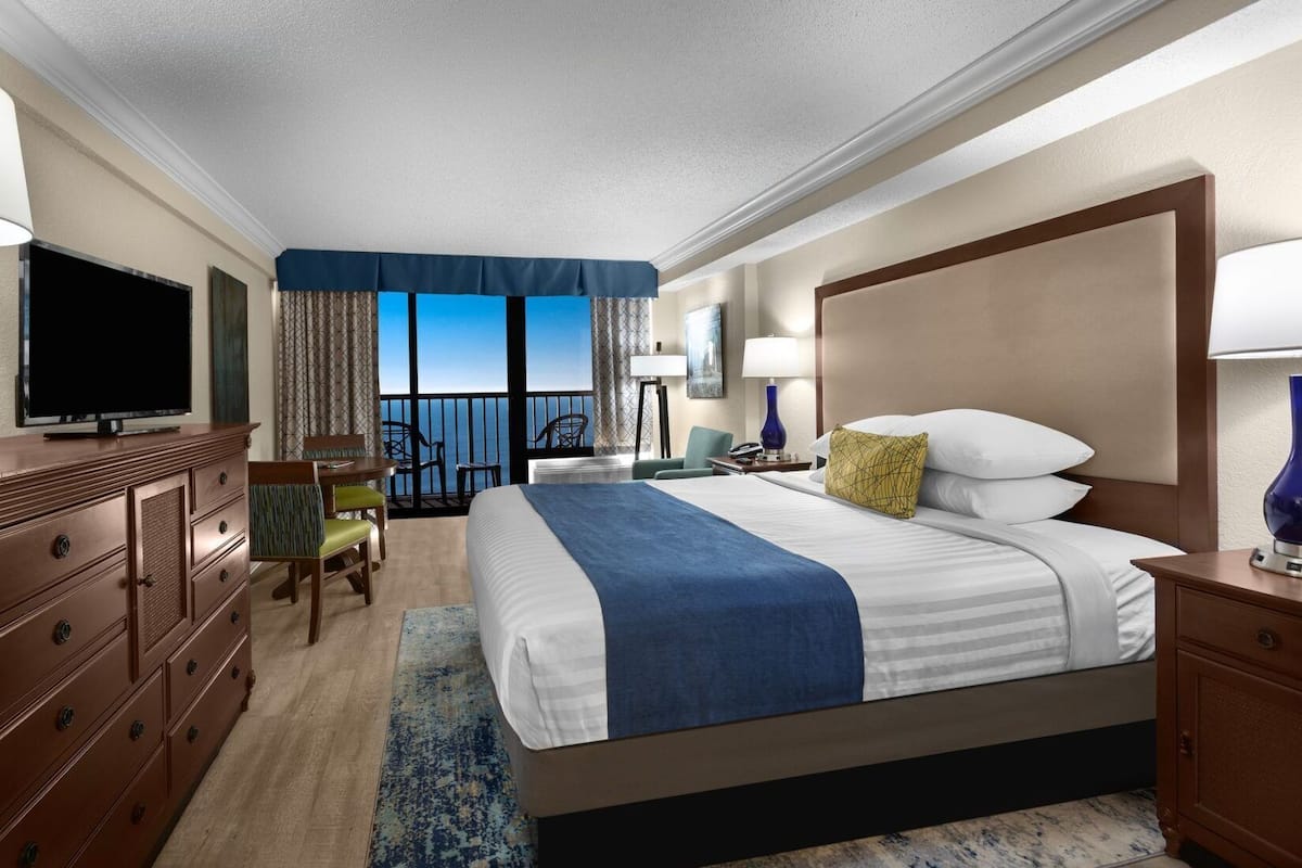 A hotel room with a large bed, a television, a desk, and a view of the ocean.