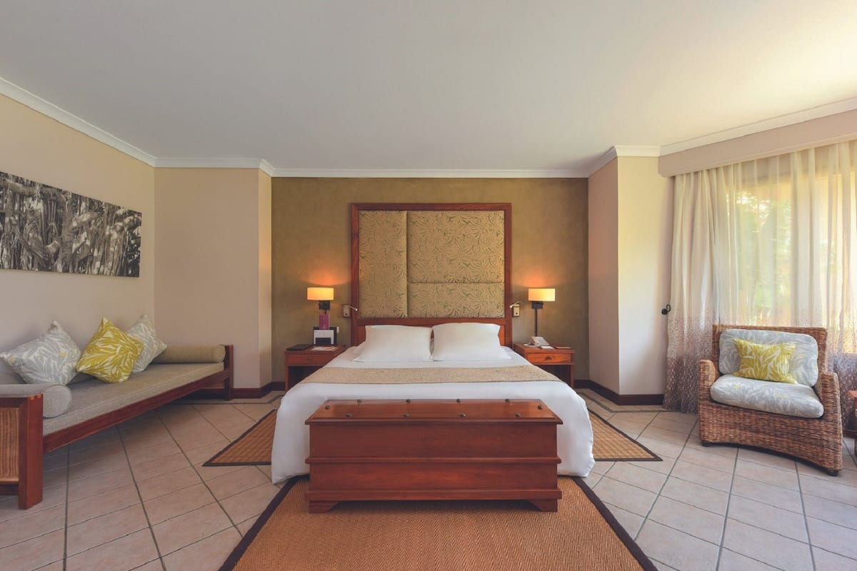 A hotel room with a large bed, bedside tables with lamps, a sofa, a chair, and a large artwork on the wall.