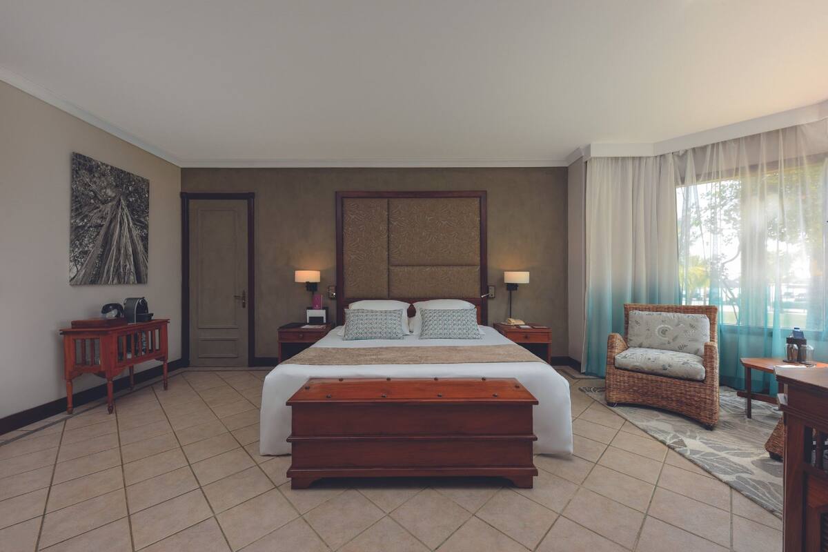 A spacious bedroom with a large bed, bedside tables, a wooden bench, a wicker chair, and a view of the outdoors through sheer curtains.