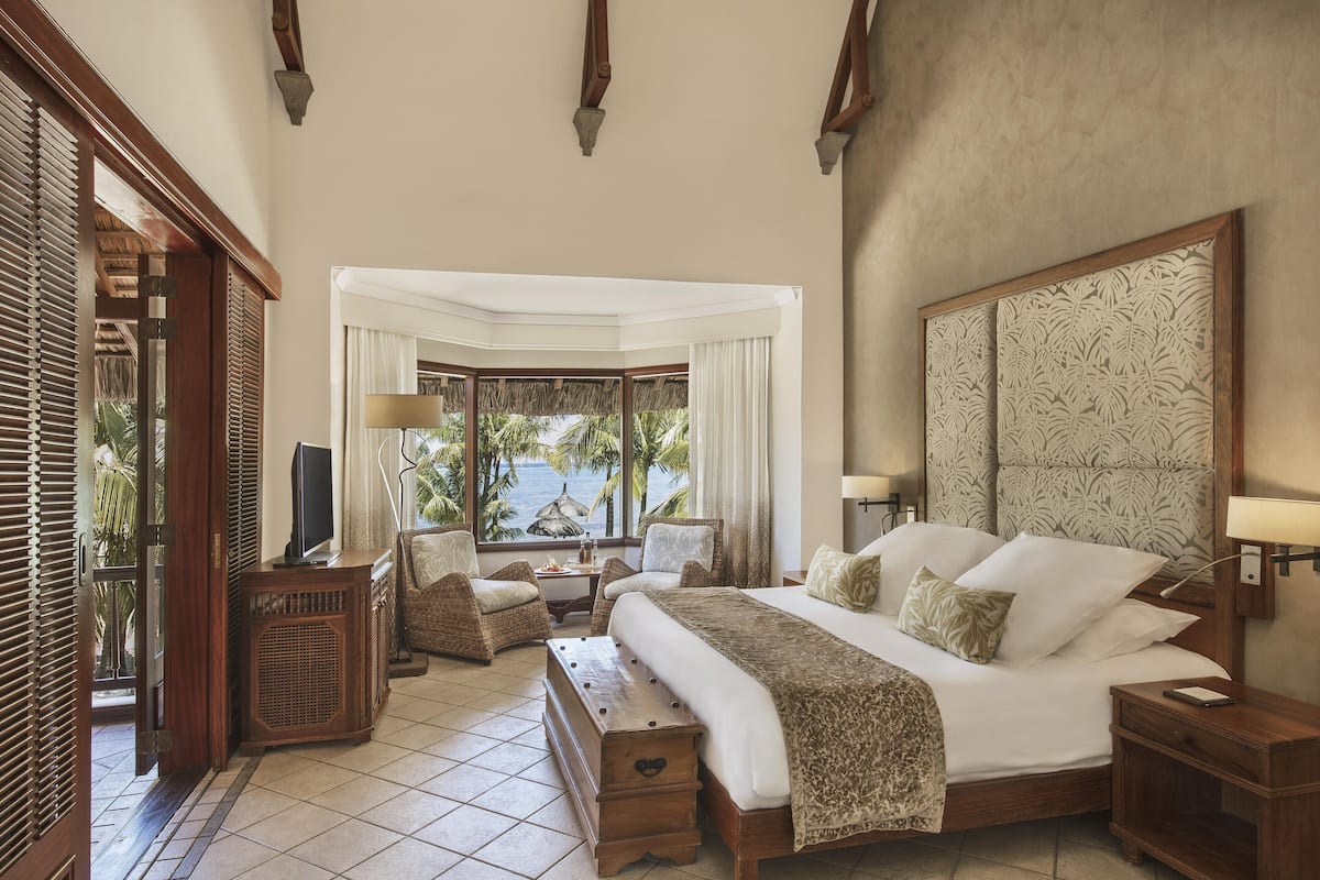 A spacious bedroom with a large bed, a sitting area with a table and chairs, and a view of palm trees and the ocean through a window.
