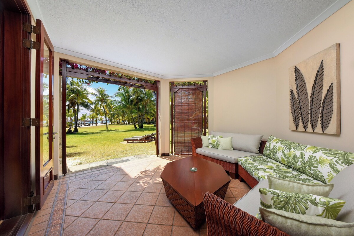 A room with a sofa, a coffee table, and a view of a garden with palm trees.