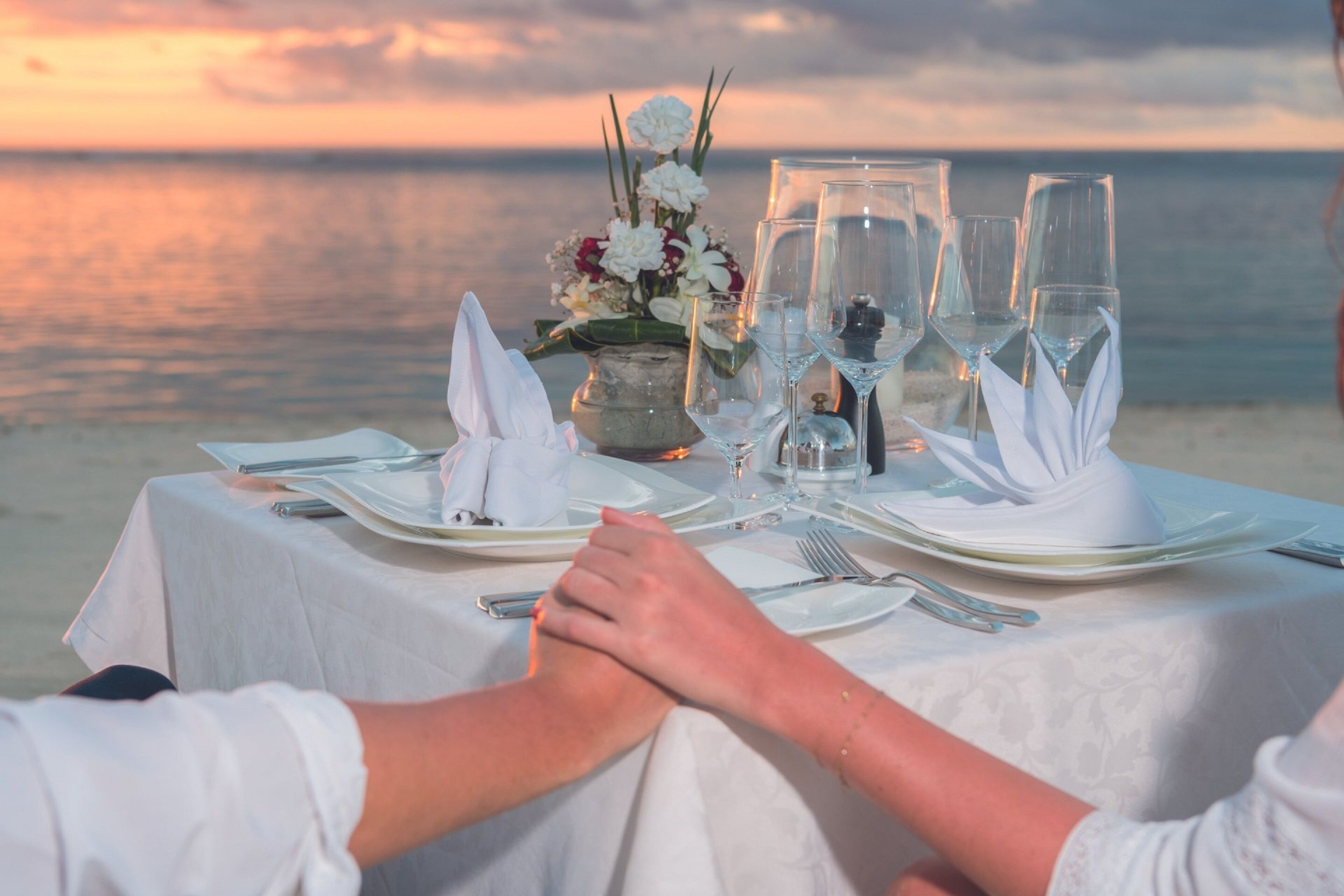 A table set for two with a vase of flowers, clear glasses, and a candle on a beach at sunset.