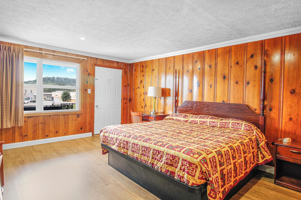 Une pièce avec un mur lambrissé en bois, un grand lit recouvert d’une couvre-lit à motifs, une table de chevet avec une lampe et une fenêtre donnant sur des bâtiments et des arbres.