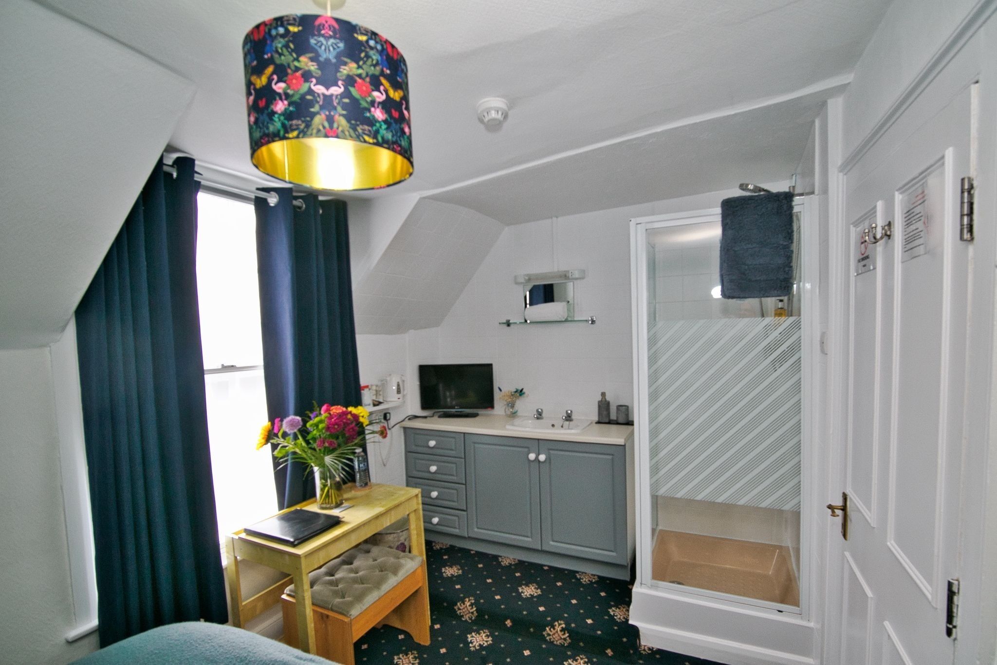 Una habitación con una cama, un escritorio con una silla, una mesita con un jarrón de flores, un televisor, un lavamanos y una ducha.