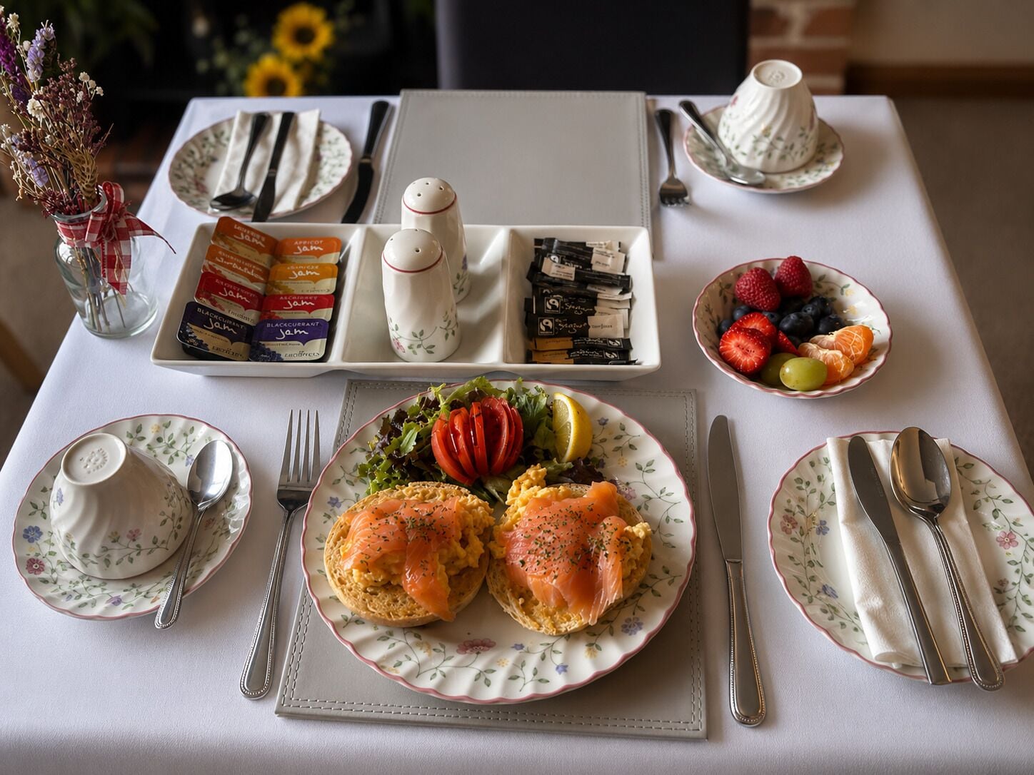 En frukostuppdukning med tallrikar fyllda med mat, en vas med blommor och ett fat med sylt.