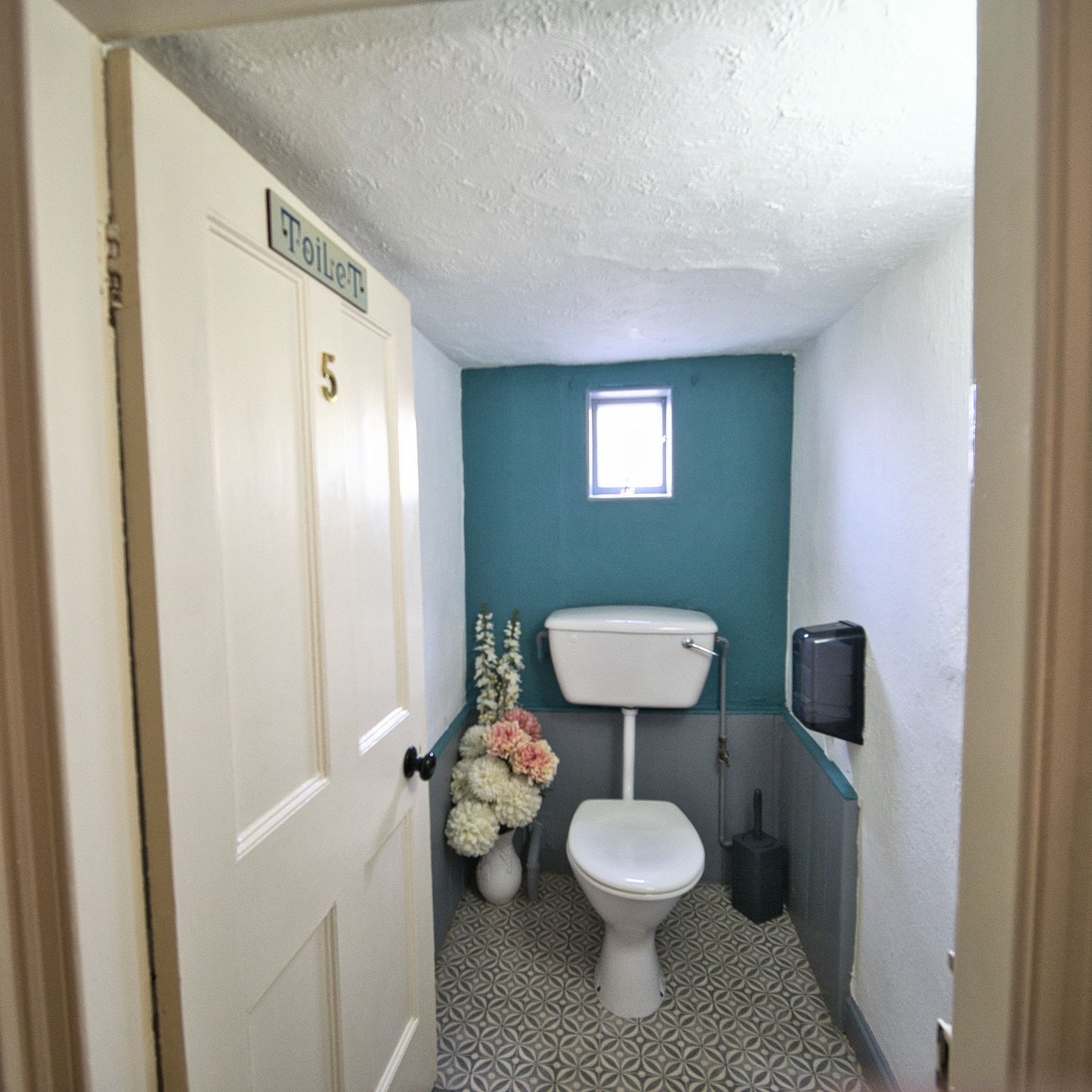 Ein Badezimmer mit Toilette, einem Fenster, einer Tür mit der Aufschrift '5' und einem Wandmülleimer.