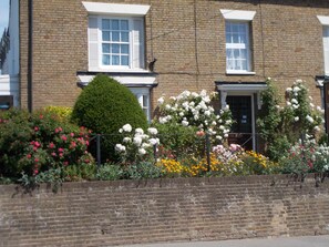 A brick house with white window shutters, a well-maintained garden with various flowers, and a neatly trimmed hedge. - The Beaumont - B&B (Sittingbourne)